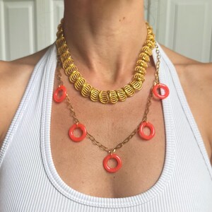May include: A layered necklace set consisting of a short, thick gold-tone chain with textured, ribbed cylindrical links and a longer, thinner gold-tone chain featuring five small, bright orange circular charms spaced evenly along the chain.