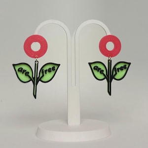 May include: A pair of statement earrings featuring a red circular top and a flower design. The flower has green leaves with the words "are free" in black. The earrings are displayed on a white stand.