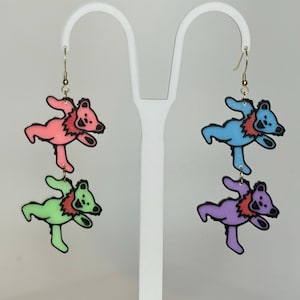 May include: Colorful bear-shaped earrings. The earrings feature dancing bears in pink, green, blue, and purple with red accents and black outlines. Each earring has a gold-colored hook.