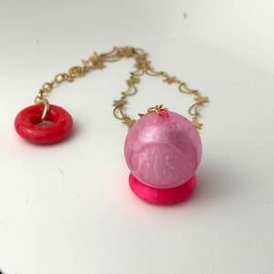 May include: A gold-toned chain featuring alternating star and crescent moon links connects a bright red ring to a spherical, pearlescent pink pendant. The pink sphere rests on a solid, vibrant hot pink circular base, creating a distinct contrast.