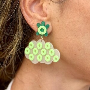 May include: A pair of unique earrings featuring a green flower stud and a cloud-shaped dangle. The cloud is white with multiple green circles. The earrings are designed to be eye-catching and add a pop of color.