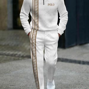 Peut inclure: Ensemble de survêtement blanc avec un motif géométrique brun sur le côté. La veste a une fermeture éclair et un col. Le pantalon a une jambe droite. Le texte "POLO" est imprimé sur la veste.