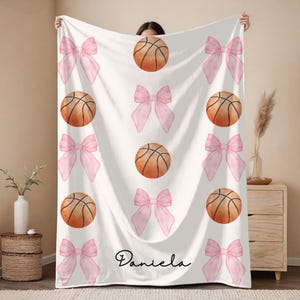 May include: White blanket featuring a pattern of pink bows and basketballs. The basketballs are orange with black lines. The name "Daniela" is written in cursive at the bottom. This blanket is perfect for a sports fan.