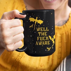 Op de afbeelding: Zwarte keramische mok met een gouden drakenontwerp en de tekst "WELL THE FUCK AWARE". De mok heeft een groot handvat. Het ontwerp bevat sterren en andere drakenillustraties. De mok wordt vastgehouden door een persoon die een gele top draagt.