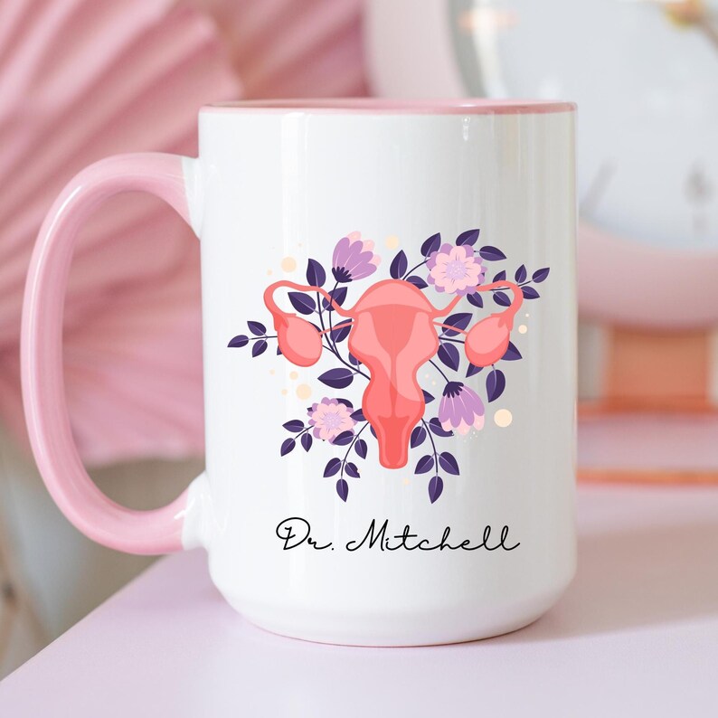 K&ouml;nnte beinhalten: Wei&szlig;er Keramikbecher mit rosa Griff und Rand. Der Becher zeigt eine stilisierte Illustration einer Geb&auml;rmutter, umgeben von lila Blumen und Bl&auml;ttern. Der Text "Dr. Mitchell" ist in schwarzer Schrift auf dem Becher geschrieben.