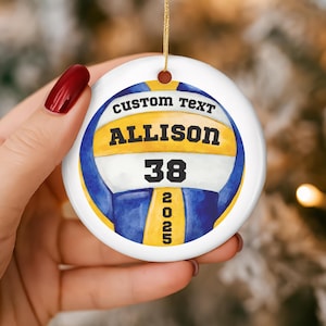 May include: A white, round ornament with a volleyball design. The ornament features the text "CUSTOM TEXT" above the name "ALLISON" and the number "38". Below the number is the year "2025". The volleyball design is yellow and blue.