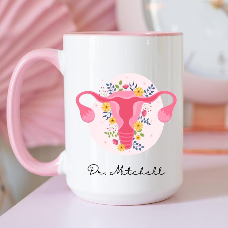 K&ouml;nnte beinhalten: Wei&szlig;e Keramiktasse mit rosa Griff und Rand. Die Tasse zeigt eine rosa und florale Illustration einer Geb&auml;rmutter. Der Text "Dr. Mitchell" ist in schwarzer Schrift auf der Tasse geschrieben.