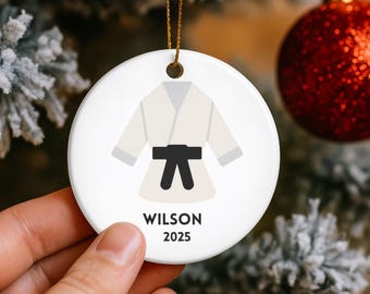 May include: White ceramic ornament with a gold string, featuring a karate uniform design with a black belt. The name "WILSON" and the year "2025" are printed below the uniform. The ornament is round and is hanging on a Christmas tree.