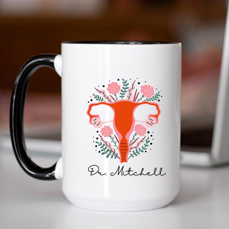K&ouml;nnte beinhalten: Wei&szlig;er Keramikbecher mit schwarzem Griff und Rand. Der Becher zeigt ein stilisiertes orangefarbenes Geb&auml;rmutterdesign, umgeben von rosa Blumen und gr&uuml;nen Bl&auml;ttern. Der Text "Dr. Mitchell" steht darunter in schwarzer Schrift.