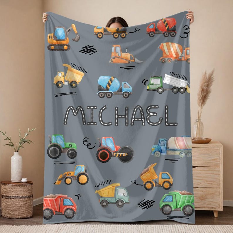 Op de afbeelding: Een grijze deken met kleurrijke illustraties van bouwmachines, waaronder graafmachines, dumper trucks en betonmixers. De naam "MICHAEL" is in witte blokletters gedrukt. De deken wordt door een persoon vastgehouden.