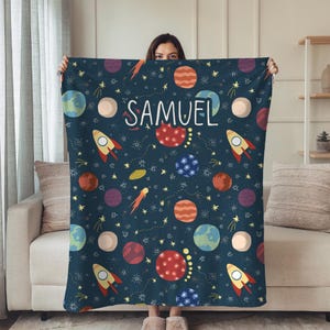 May include: A navy blue blanket featuring a colorful space-themed pattern with planets, rockets, and stars. The name "SAMUEL" is written in white script across the blanket. The blanket is held up in front of a person.