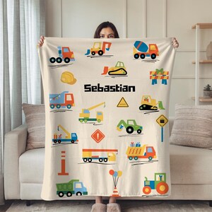 以下が含まれることがあります： ベージュ色のブランケット。トラック、クレーン、トラクターなど、建設車両のカラフルなイラストが描かれています。「Sebastian」という名前が黒いブロック体で印刷されています。人がブランケットを持っています。
