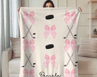May include: Cream-colored blanket featuring a pattern of pink bows, hockey sticks, and pucks. The blanket has the name "Daniela" in a script font. The design is repeated across the blanket.