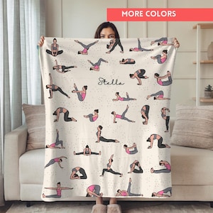 Könnte beinhalten: Eine cremefarbene Decke mit Illustrationen von Yoga-Posen in Schwarz, Grau und Rosa. Die Decke trägt den Namen "Stella" in Schreibschrift. Die Worte "MORE COLORS" stehen in einem roten Rechteck oben rechts.