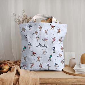 May include: A light blue tote bag featuring a pattern of ballet dancers in various poses. The bag has white handles and the name "Mady" in cursive script. The dancers are in different colors, and the bag is sitting on a wooden surface.