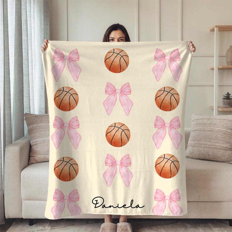 May include: A cream-colored blanket featuring a pattern of pink bows and basketballs. The basketballs are orange with black lines. The blanket has the name "Daniela" in a cursive font at the bottom.