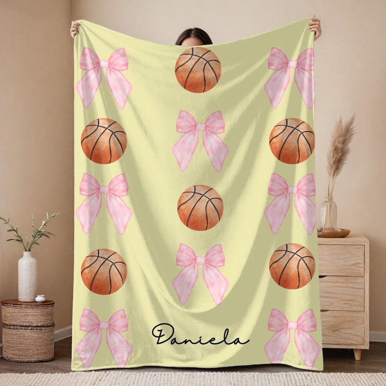 May include: A light yellow blanket featuring a pattern of pink bows and basketballs. The basketballs are orange with black lines. The name "Daniela" is written in black script at the bottom.