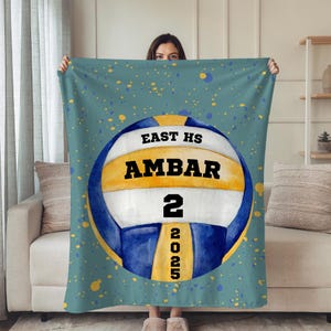 Pode incluir: Um cobertor turquesa com um design de voleibol. O texto na bola de voleibol diz "EAST HS AMBAR 2 2025." A bola de voleibol é branca, amarela e azul, sobre um fundo turquesa com salpicos de tinta amarelos e azuis. O cobertor é segurado.