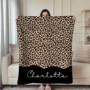 Puede incluir: Una manta con estampado de leopardo en tonos beige y marrón, con un borde negro que lleva el nombre "Charlotte" en escritura blanca. La manta está siendo sostenida por una persona. Las dimensiones de la manta no son visibles.