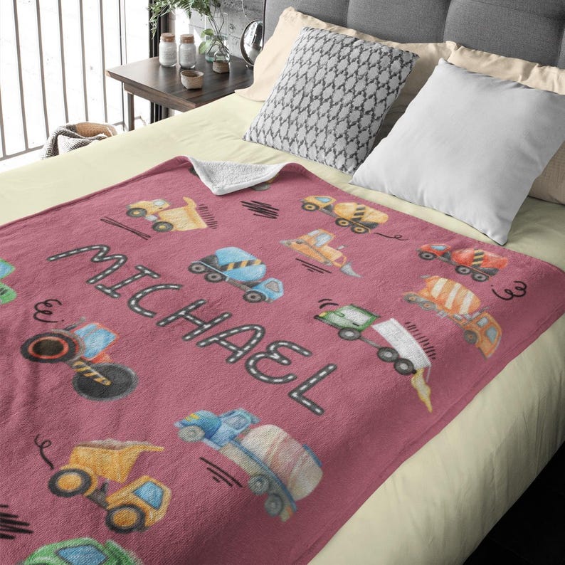 Op de afbeelding: Een stoffige roze deken met een patroon van gele, blauwe en oranje bouwmachines. De naam "MICHAEL" staat op de deken. De deken ligt op een bed met kussens.