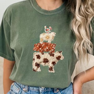 May include: Olive green t-shirt featuring a stacked animal design. The design includes a cow, a pig, and a chicken, each with a unique floral pattern. The cow has brown spots, the pig has a brown floral pattern, and the chicken has a pink floral pattern.