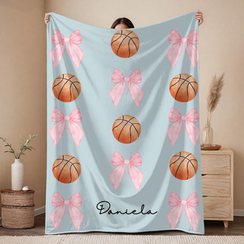May include: A light blue blanket featuring a pattern of pink bows and basketballs. The basketballs are brown with black lines. The name "Daniela" is written in cursive at the bottom of the blanket.