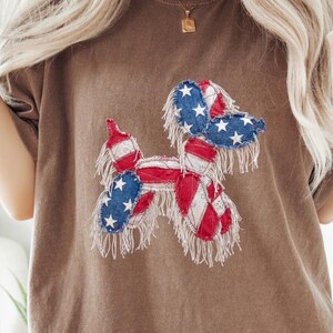 Op de afbeelding: Een bruin t-shirt met een patriottisch hondenontwerp. De hond is gemaakt van rode, witte en blauwe patches, die lijken op de Amerikaanse vlag. De hond heeft witte franjes rond de randen.