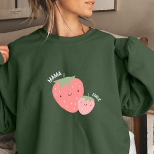 Peut inclure: Sweat-shirt vert foncé avec un motif de fraise. Le mot "MAMA" est au-dessus d'une grosse fraise, et le nom "EMILIE" est au-dessus d'une petite fraise. Les fraises sont roses avec des sommets verts et des visages souriants.