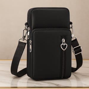 May include: A black crossbody bag with a rectangular shape and a long, adjustable strap. The bag features a zippered main compartment and a smaller front pocket. A heart-shaped charm hangs from the zipper pull. The bag is made of a smooth, matte material.