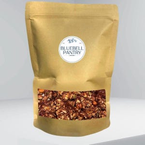 May include: A tan paper stand-up pouch with a clear rectangular window displaying chocolate granola with nuts. A circular white label on the front features a blue bell icon and the text BLUEBELL PANTRY with EST. 2024 printed in small font.