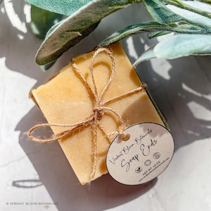 Puede incluir: Una pastilla de jabón rectangular de color amarillo claro, atada con hilo y una pequeña etiqueta. La etiqueta dice "Verdant Bloom Botanicals Soap Ends". El peso neto es de 128 gramos.