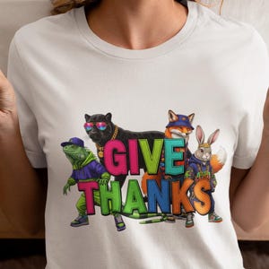 May include: White t-shirt with a colorful graphic that reads "GIVE THANKS". The graphic features a frog, panther, fox, and rabbit wearing sunglasses and clothing. The shirt is a crew neck and has short sleeves.
