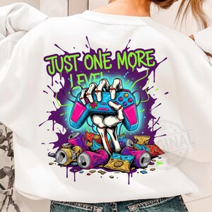 Könnte beinhalten: Weißes Sweatshirt mit einem grafischen Design einer Skeletthand, die einen blau-rosa Videospiel-Controller hält. Der Text "JUST ONE MORE LEVEL" befindet sich über dem Controller, mit verstreuten Dosen und Snackverpackungen.