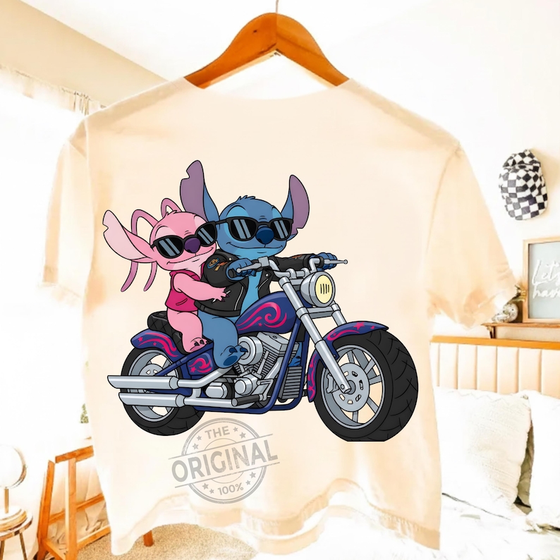 May include: A light-colored t-shirt featuring a cartoon image of two characters riding a motorcycle. The blue character is driving, wearing a black jacket and sunglasses. The pink character is a passenger, also wearing sunglasses. The motorcycle is purple and blue.