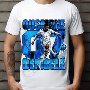 May include: White t-shirt featuring a graphic of soccer player Ousmane Demb&eacute;l&eacute; in action, kicking a soccer ball. The design includes the player's name and jersey number in blue, with a French flag detail.