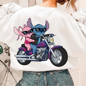 May include: White sweatshirt featuring a cartoon illustration of two characters riding a motorcycle. The blue character wears a black leather jacket and sunglasses, while the pink character wears sunglasses and a pink top. The motorcycle is blue with pink accents.