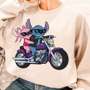 May include: A beige sweatshirt featuring a cartoon illustration of two aliens riding a motorcycle. The blue alien is driving, wearing sunglasses and a black leather jacket. The pink alien is a passenger, also wearing sunglasses. The motorcycle is purple and blue.