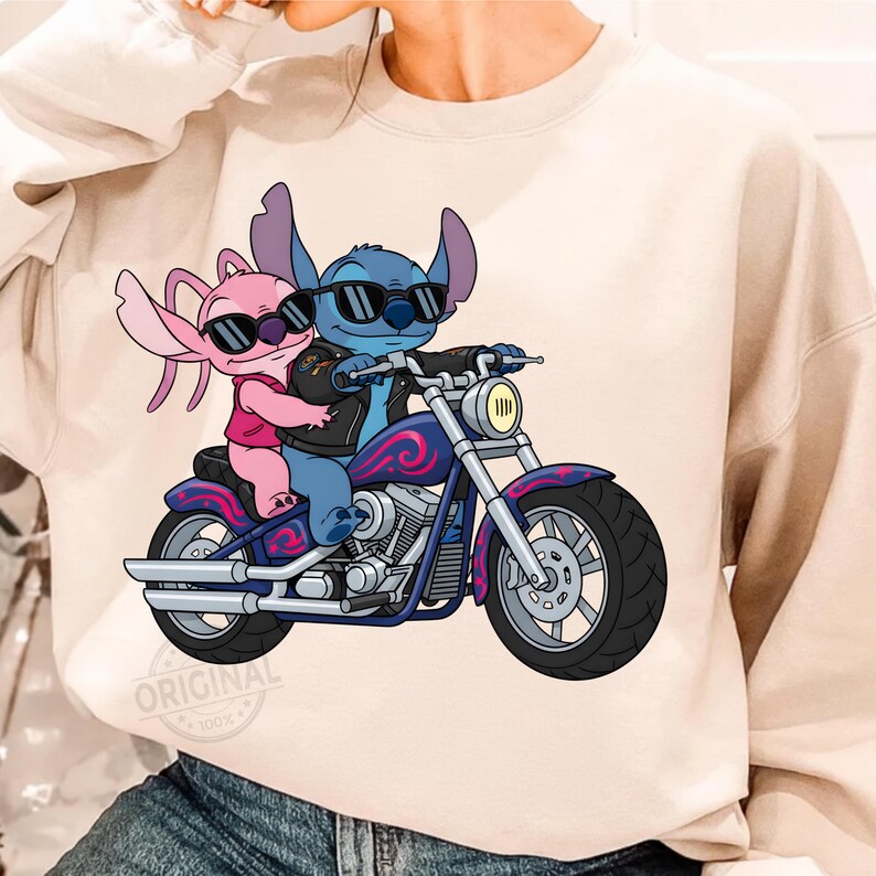 May include: A beige sweatshirt featuring a cartoon illustration of two aliens riding a motorcycle. The blue alien is driving, wearing sunglasses and a black leather jacket. The pink alien is a passenger, also wearing sunglasses. The motorcycle is purple and blue.