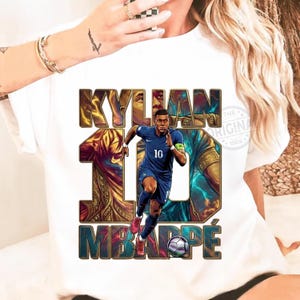 May include: White t-shirt featuring a graphic of a soccer player in a blue uniform, with the name "KYLIAN MBAPP&Eacute;" in bold letters. The number "10" is visible on the player's jersey. A soccer ball is also included in the design.