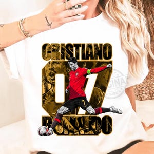 May include: White t-shirt featuring a graphic of Cristiano Ronaldo in a red soccer uniform, kicking a soccer ball. The design includes the name "CRISTIANO" and "RONALDO" in gold and black, with the number "07".