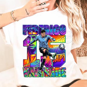 May include: White t-shirt featuring a colorful graphic of a soccer player kicking a ball. The design includes the name "FEDERICO VALVERDE" and the number "15" in vibrant, stylized lettering. The player is wearing a light blue jersey.