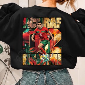 May include: Black sweatshirt featuring a graphic of a soccer player in a red and green uniform, with the name "ACHRAF" and the number "02" in large letters. The design includes the word "MAXIM" and a soccer ball.