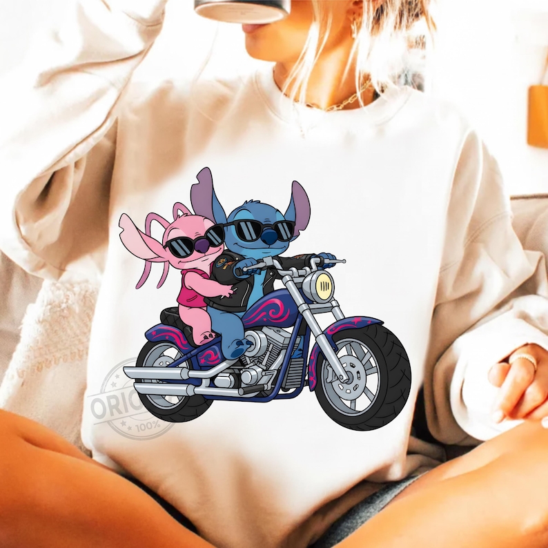 May include: White sweatshirt featuring a cartoon illustration of two aliens riding a motorcycle. The blue alien is wearing a black jacket and sunglasses, while the pink alien is wearing a pink shirt and sunglasses. The motorcycle is purple and blue.