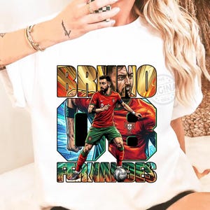 May include: White t-shirt featuring a graphic of a soccer player in a red and green uniform, with the name "BRUNO FERNANDES" and the number "8" in bold text. The design includes a portrait of the player.