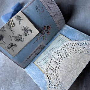 May include: Open journal with light blue pages, floral illustrations, and handwritten script. A vintage-style paper with a floral illustration is held in place with a paper clip. A white lace doily is visible.
