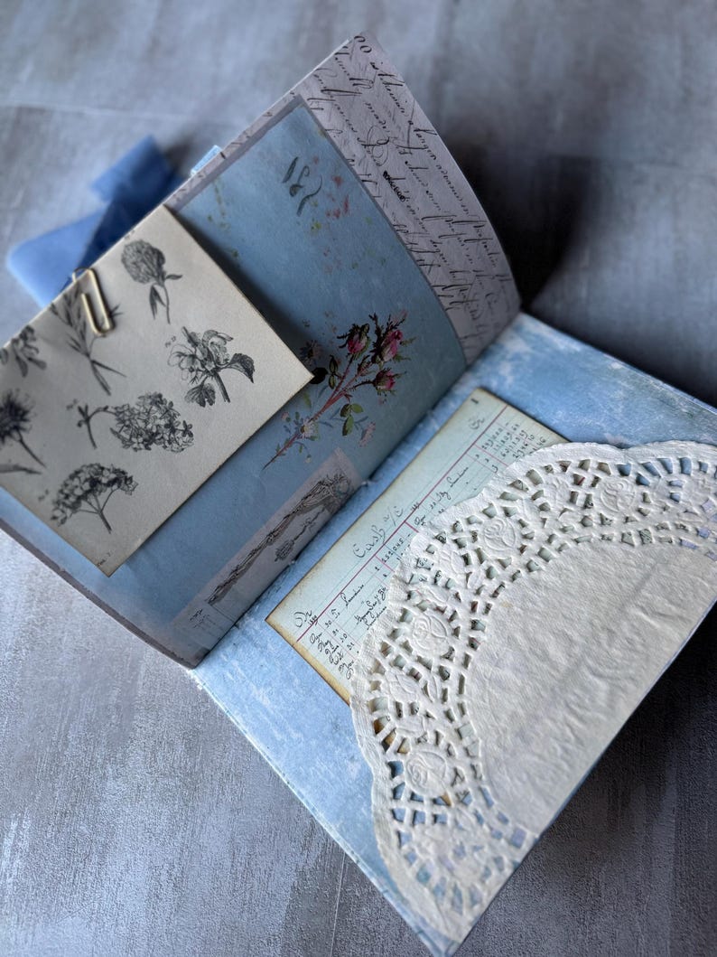 May include: Open journal with light blue pages, floral illustrations, and handwritten script. A vintage-style paper with a floral illustration is held in place with a paper clip. A white lace doily is visible.