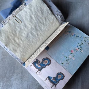 May include: Open journal with aged paper pages, secured with a silver paperclip. The journal features blue floral patterned covers and a light blue ribbon. Illustrations of blue chairs with floral details are visible.
