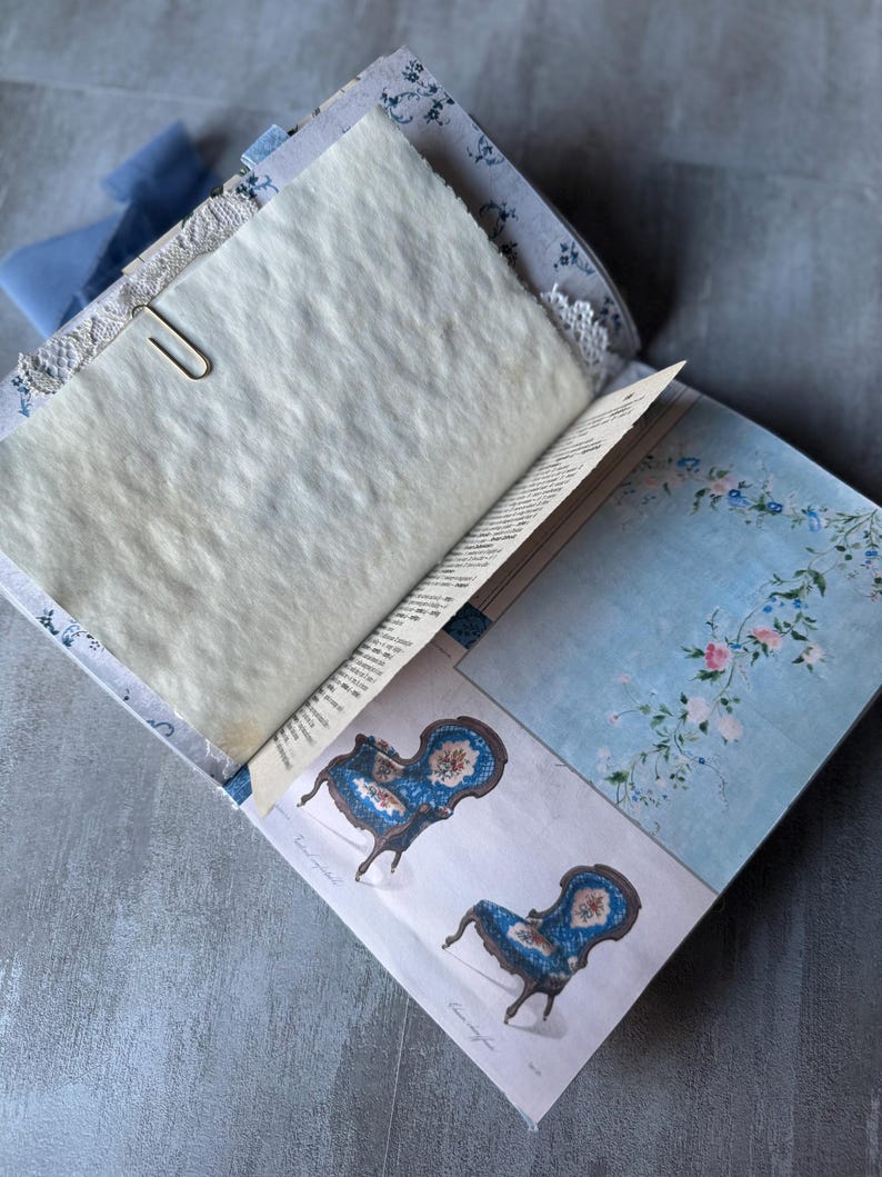 May include: Open journal with aged paper pages, secured with a silver paperclip. The journal features blue floral patterned covers and a light blue ribbon. Illustrations of blue chairs with floral details are visible.
