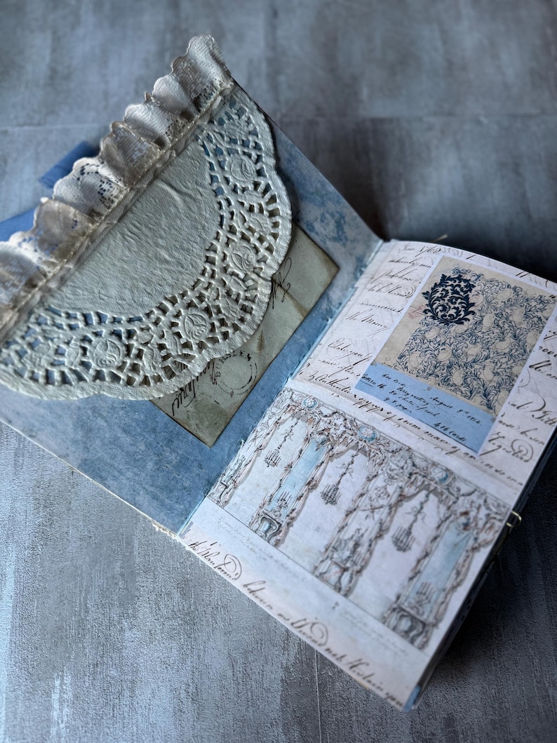 May include: Open journal with blue and white patterned pages, featuring a lace doily and handwritten script. The journal has a vintage aesthetic, with a delicate, layered design. The pages display architectural drawings and decorative elements.