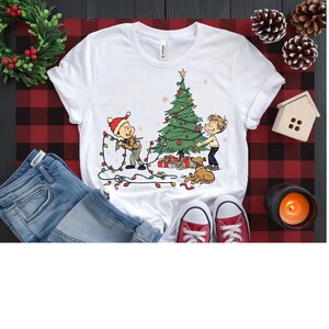 Peut inclure: Un t-shirt blanc à manches courtes orné d'une illustration graphique de deux personnages de dessin animé décorant un sapin de Noël avec des guirlandes lumineuses, un petit chien brun et des cadeaux au pied de l'arbre.